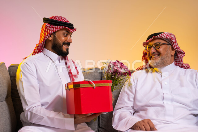 مفهوم المحبة والألفة بين أفراد العائلة، ايماءات البهجة والسرور، بورتريه مقرب لرجل عربي خليجي سعودي يرتدي الزي التقليدي والشماغ يقوم بالنظر الى ابنه بحب، شاب يقدم هدية وباقة ورد لوالده بمناسبة عيد ميلاده، الاحتفال بالأعياد والمناسبات، خلفية ملونة