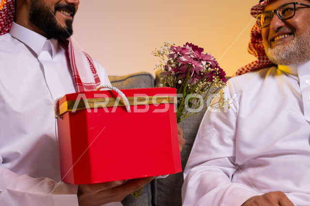 المحبة والألفة بين أفراد العائلة، ايماءات البهجة والسرور، بورتريه مقرب لرجل عربي خليجي سعودي يرتدي الزي التقليدي والشماغ يقوم بالنظر الى ابنه بحب، شاب يقدم هدية وباقة ورد لوالده بمناسبة عيد ميلاده، الاحتفال بالأعياد والمناسبات، خلفية ملونة