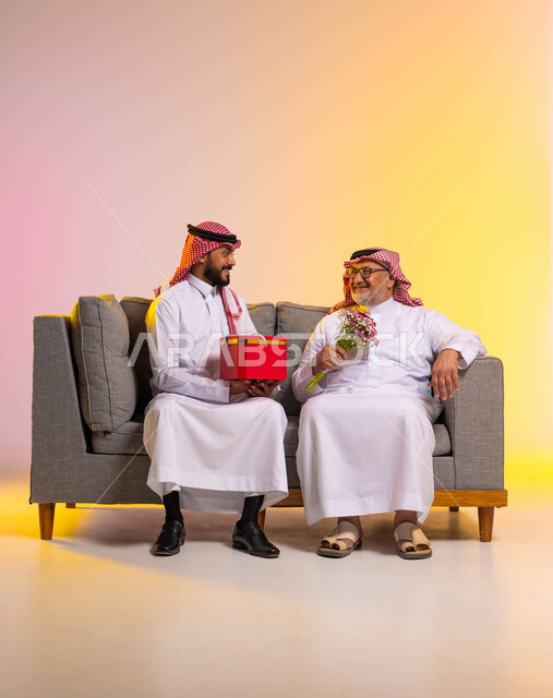 الاحتفال بالأعياد والمناسبات، ايماءات البهجة والسرور، بورتريه لرجل عربي خليجي سعودي يرتدي الزي التقليدي والشماغ يحمل بيده باقة ورد ينظر الى ابنه بحب، شاب يقدم هدية لوالده بمناسبة عيد ميلاده، الألفة والمحبة بين أفراد العائلة، الطول الكامل للجسم، خلفية ملونة