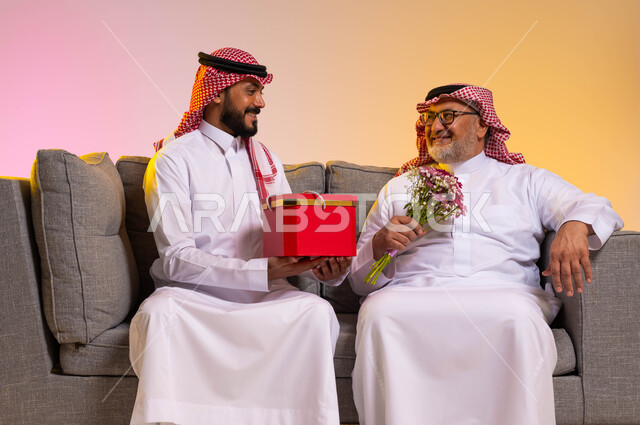ايماءات البهجة والسرور، بورتريه لرجل عربي خليجي سعودي يرتدي الزي التقليدي والشماغ يحمل بيده باقة ورد يقوم بالنظر الى ابنه بحب، شاب يقدم هدية لوالده بمناسبة عيد ميلاده، الألفة والمحبة بين أفراد العائلة، الاحتفال بالأعياد والمناسبات، خلفية ملونة