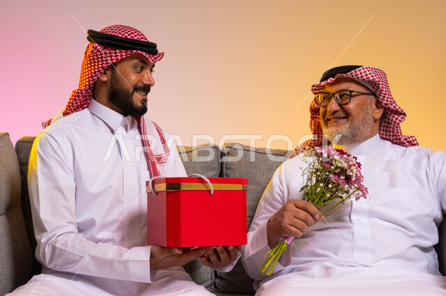 الألفة والمحبة بين أفراد العائلة، ايماءات البهجة والسرور، بورتريه مقرب لرجل عربي خليجي سعودي يرتدي الزي التقليدي والشماغ يحمل بيده باقة ورد يقوم بالنظر الى ابنه بحب، شاب يقدم هدية لوالده بمناسبة عيد ميلاده، الاحتفال بالأعياد والمناسبات، خلفية ملونة