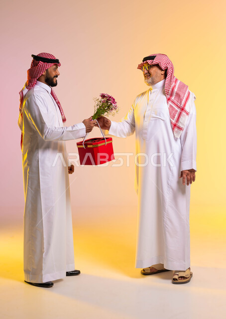 ايماءات تدل على السعادة والسرور وقبول الهدية، مشاركة وتقديم الهدايا في الأعياد والمناسبات السعيدة، بورتريه من الجانب لشاب عربي خليجي سعودي يرتدي الثوب التقليدي والشماغ يقوم باهداء والده صندوق أحمر وباقة ورد، حب وإحترام الآباء، الطول الكامل للجسم، خلفية ملونة