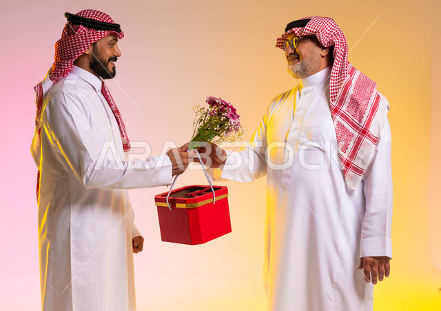 مشاركة وتقديم الهدايا في الأعياد والمناسبات السعيدة، بورتريه من الجانب لشاب عربي خليجي سعودي يرتدي الثوب التقليدي والشماغ يقوم باهداء والده صندوق أحمر وباقة ورد، ايماءات تدل على السعادة والسرور وقبول الهدية، حب وإحترام الآباء، خلفية ملونة