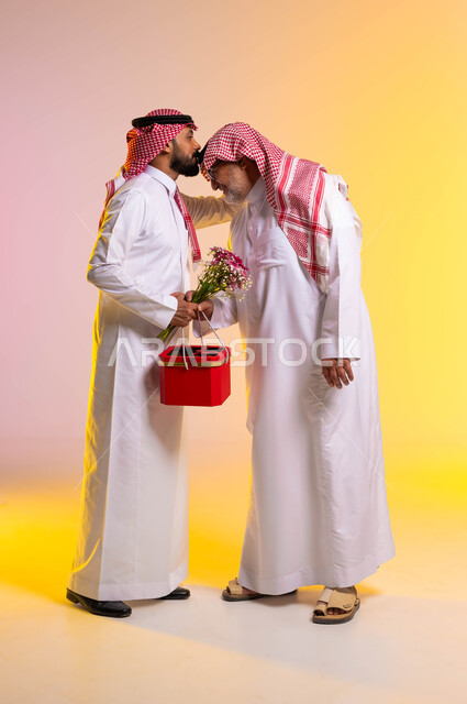 إرتداء الأزياء الشعبية التقليدية في الأعياد والمناسبات، مفهوم التربية الصالحة للأبناء، بورتريه من الجانب لشاب عربي خليجي سعودي يحمل بيده باقة ورد وصندوق أحمر يقوم بتقبيل رأس والده، إحترام وتقدير الآباء والإحسان إليهم، الطول الكامل للجسم، خلفية ملونة