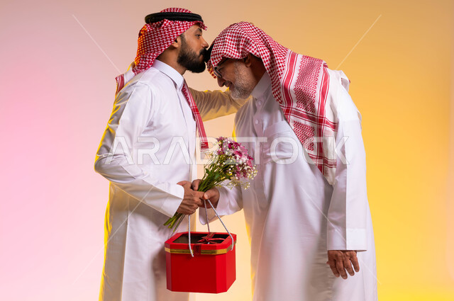 مفهوم التربية الصالحة للأبناء، بورتريه من الجانب لشاب عربي خليجي سعودي يحمل بيده باقة ورد وصندوق أحمر يقوم بتقبيل رأس والده، إرتداء الأزياء الشعبية التقليدية في الأعياد والمناسبات، إحترام وتقدير الآباء والإحسان إليهم، خلفية ملونة