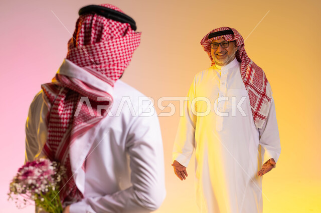 مفهوم إحترام وتقدير الآباء وادخال الفرحة والسرور عليهم، إهداء الهدايا في الأعياد والمناسبات السعيدة، بورتريه مقرب من الخلف لشاب عربي خليجي سعودي يرتدي الثوب التقليدي والشماغ يخبئ خلفه باقة ورد، مفاجأة الوالد في عيد ميلاده، خلفية ملونة