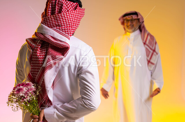 مفهوم تقدير وإحترام الآباء وادخال الفرحة والسرور عليهم، إهداء الهدايا في الأعياد والمناسبات السعيدة، بورتريه مقرب من الخلف لشاب عربي خليجي سعودي يرتدي الثوب التقليدي والشماغ يخبئ خلفه باقة ورد، مفاجأة الوالد في عيد ميلاده، خلفية ملونة