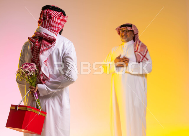 مفهوم أهمية الأسرة، تقديم الهدايا للاهل في المناسبات والأعياد، الإحسان الى الآباء ورد الجميل، ايماءات الدهشة والسرور، بورتريه من الخلف لشاب عربي خليجي سعودي يرتدي الثوب التقليدي والشماغ يحمل بيده باقة من الورد وصندوق أحمر يقوم بمفاجأة والده بحب، خلفية ملونة