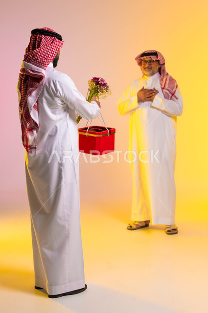 تقديم الهدايا للاهل في المناسبات والأعياد، الإحسان الى الآباء و رد الجميل، ايماءات الدهشة والسرور، مفهوم أهمية الأسرة، بورتريه من الخلف لشاب عربي خليجي سعودي يرتدي الثوب التقليدي والشماغ يحمل بيده باقة من الورد وصندوق أحمر، صورة لكامل الجسم، خلفية ملونة