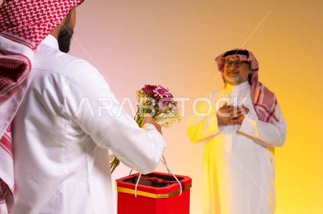 ايماءات الدهشة والسرور، الإحسان الى الآباء ورد الجميل، تقديم الهدايا للاهل في المناسبات والأعياد، مفهوم أهمية الأسرة، بورتريه من الخلف لشاب عربي خليجي سعودي يرتدي الثوب التقليدي والشماغ يحمل بيده باقة من الورد وصندوق أحمر، خلفية ملونة