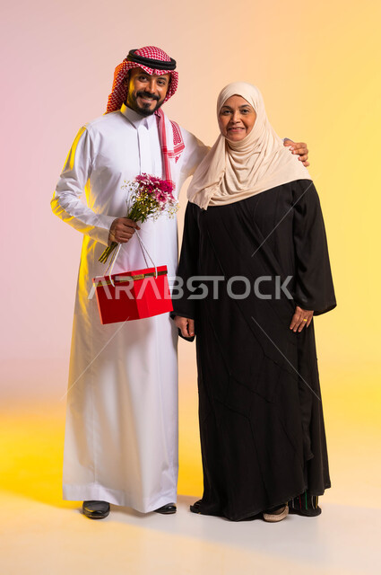 مفهوم أهمية الأسرة، تبادل المودة والمحبة بين أفراد العائلة، بورتريه لشاب عربي خليجي سعودي يرتدي الزي التقليدي يقف بجانب والدته يحمل بيده باقة من الأزهار، النظر الى الكاميرا بايماءات السعادة والسرور، تقديم الهدايا في الأعياد، صورة الجسم بالكامل، خلفية ملونة