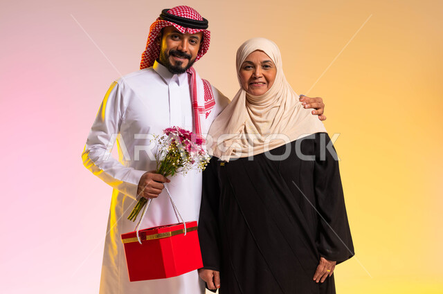 تقديم الهدايا في الأعياد والمناسبات، النظر الى الكاميرا بايماءات البهجة والسرور، تبادل المودة والمحبة بين أفراد العائلة، بورتريه لشاب عربي خليجي سعودي يرتدي الزي التقليدي يقف بجانب والدته يحمل بيده باقة من الورد، مفهوم أهمية الأسرة، خلفية ملونة