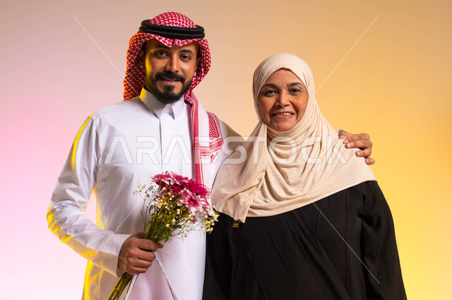 النظر الى الكاميرا بايماءات البهجة والسرور، تبادل المودة والمحبة بين أفراد العائلة، بورتريه لشاب عربي خليجي سعودي يرتدي الزي التقليدي يقف بجانب والدته يحمل بيده باقة من الورد، تقديم الهدايا في الأعياد والمناسبات، مفهوم أهمية الأسرة، خلفية ملونة