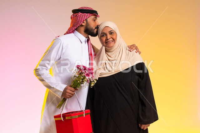 احترام الأم والبر بها، تبادل المودة والمحبة بين أفراد الأسرة، بورتريه لشاب عربي خليجي سعودي يرتدي الزي التقليدي يقوم بتقبيل رأس والدته، أجواء عائلية سعيدة، ايماءات السعادة والسرور، تقديم الهدايا والأزهار في الأعياد والمناسبات، خلفية ملونة