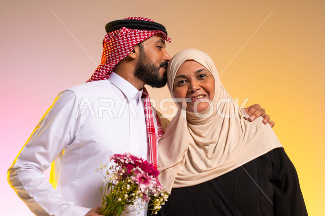 مفهوم البر بالوالدين، تبادل المودة والمحبة بين أفراد الأسرة، بورتريه مقرب لشاب عربي خليجي سعودي يرتدي الزي التقليدي يقوم بتقبيل رأس والدته، أجواء عائلية سعيدة، ايماءات السعادة والسرور، تقديم الهدايا والأزهار في الأعياد والمناسبات، خلفية ملونة