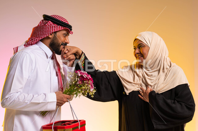 التعبير عن الحب والاحترام للأم، مفهوم الترابط والتماسك الأسري، تقديم الهدايا والورد للاهل في المناسبات والأعياد، بورتريه لشاب عربي خليجي سعودي يرتدي الزي التقليدي يقوم بتقبيل يد والدته، ايماءات البهجة والسرور، خلفية ملونة