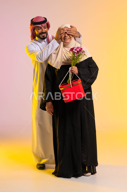 التربية الصالحة والبر بالوالدين، تقديم الهدايا والأزهار في المناسبات والأعياد، بورتريه لشاب عربي خليجي سعودي يرتدي الثوب التقليدي والشماغ يغمض عيني والدته بيديه، مفاجأة الأم وإدخال السرور عليها، صورة لكامل الجسم، خلفية ملونة