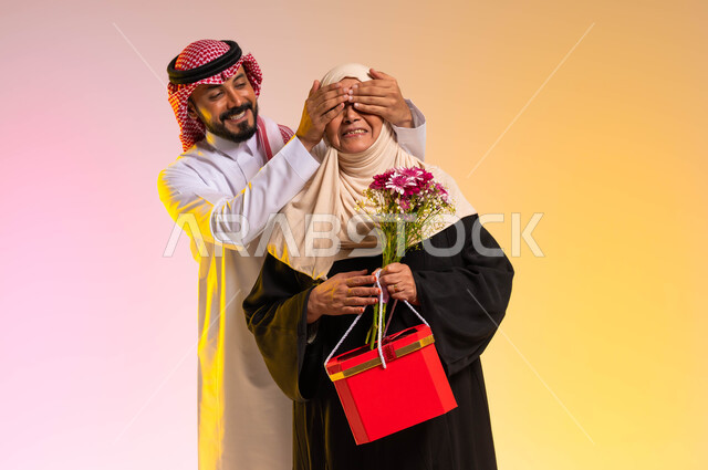 Providing gifts and flowers on holidays and occasions, the concept of respecting and appreciating the mother and being kind to her, family bonding and cohesion, a portrait of a Saudi Gulf Arab young man wearing the traditional dress and shemagh, covering his mother’s eyes with his hands, surprising the mother and making her happy, a colorful background.