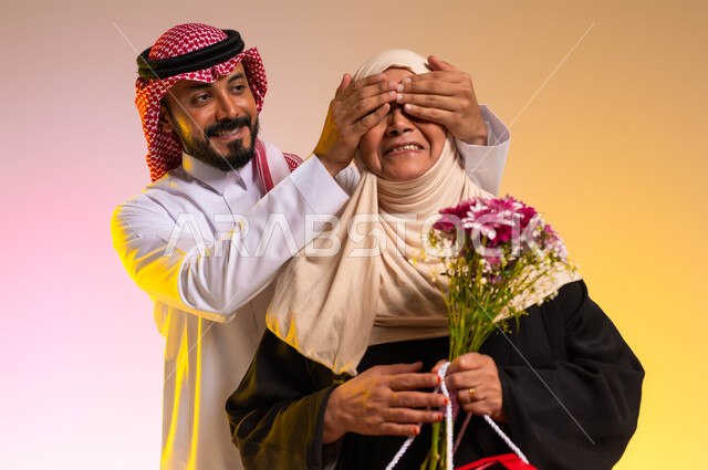 مفاجأة الأم وإدخال السرور عليها، مفهوم احترام وتقدير الوالدة والإحسان إليها، تقديم الهدايا والأزهار في الأعياد والمناسبات، بورتريه مقرب لشاب عربي خليجي سعودي يرتدي الثوب التقليدي والشماغ يغمض عيني والدته بيديه، خلفية ملونة