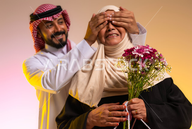 مفهوم احترام وتقدير الوالدة والإحسان إليها، تقديم الهدايا والأزهار في الأعياد والمناسبات، بورتريه مقرب لشاب عربي خليجي سعودي يرتدي الثوب التقليدي والشماغ يغمض عيني والدته بيديه، مفاجأة الأم وإدخال السرور عليها، خلفية ملونة