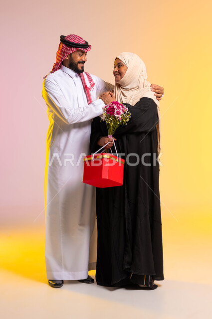 التربية الصالحة للأولاد، تبادل المحبة والمودة بين الآباء والأبناء، الإحسان الى الوالدة ورد الجميل، تقديم الهدايا والأزهار للاهل في الأعياد والمناسبات، بورتريه لشاب عربي خليجي سعودي يرتدي الثوب التقليدي والشماغ يقف بجانب والدته، صورة الجسم بالكامل، خلفية ملونة