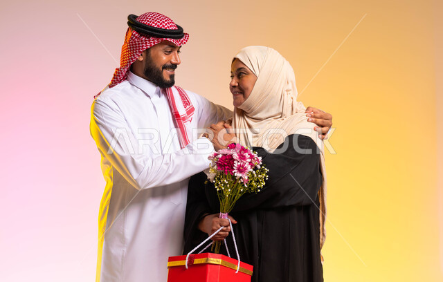 تبادل المحبة والمودة بين الآباء والأبناء، الإحسان الى الوالدة و رد الجميل، تقديم الهدايا والزهور للاهل في الأعياد والمناسبات، بورتريه لشاب عربي خليجي سعودي يرتدي الثوب التقليدي والشماغ يقف بجانب والدته، التربية الصالحة والبر بالأم، خلفية ملونة