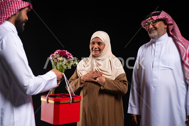 إدخال البهجة والسرور على قلب الأم، الإحسان الى الآباء والبر بهما، بورتريه مقرب لعائلة عربيية خليجية سعودية ترتدي الزي التقليدي تحتفل في الأعياد والمناسبات السعيدة، شاب يقدم هدية بصندوق أحمر وباقة ورد لوالديه، مفهوم الترابط الأسري، خلفية سوداء