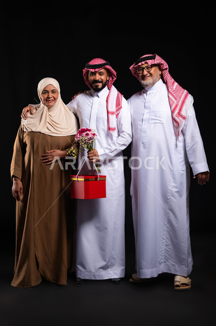 تقديم الهدايا للاهل في الأعياد والمناسبات، أجواء عائلية مليئة بالسعادة، المحبة والترابط الأسري، تبادل المودة بين أفراد الأسرة، بورتريه لعائلة عربية خليجية سعودية ترتدي الزي التقليدي تنظر إلى الكاميرا بإيماءات البهجة والسرور، صورة لكامل الجسم، خلفية سوداء