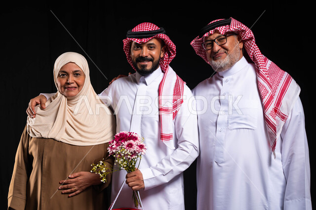 A family atmosphere full of happiness, love and family bonding, the exchange of affection between family members, a close-up portrait of a Saudi Gulf Arab family wearing traditional clothing looking at the camera with gestures of joy and happiness, presenting gifts and flowers to the family on holidays and occasions, a black background.