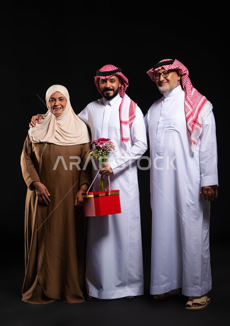 المحبة والترابط الأسري، تبادل المودة بين أفراد الأسرة، بورتريه لعائلة عربية خليجية سعودية ترتدي الزي التقليدي تنظر إلى الكاميرا بإيماءات السرور، أجواء عائلية مليئة بالحب، تقديم الهدايا والأزهار للأهل في الأعياد والمناسبات، صورة لكامل الجسم، خلفية سوداء