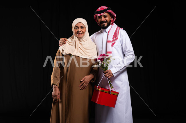 تقديم الهدايا في الأعياد والمناسبات، النظر الى الكاميرا بايماءات السعادة والسرور، تبادل المودة والمحبة بين أفراد الأسرة، بورتريه لشاب عربي خليجي سعودي يرتدي الزي التقليدي يضع يده على كتف والدته يحمل بيده هدية في صندوق أحمر وباقة ورد، خلفية سوداء