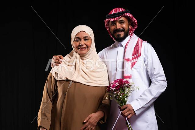 النظر الى الكاميرا بايماءات البهجة والسرور، تبادل المودة والمحبة بين أفراد العائلة، بورتريه لشاب عربي خليجي سعودي يرتدي الزي التقليدي يقف بجانب والدته يحمل بيده باقة من الورد، تقديم الهدايا في الأعياد والمناسبات، مفهوم البر بالوالدين والإحسان إليهما، خلفية سوداء
