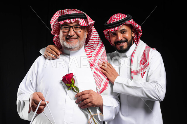 Appreciating and respecting parents and being kind to them, offering gifts and flowers on holidays and occasions, a close-up portrait of an elderly Saudi Gulf Arab man wearing the traditional dress and shemagh, holding a gift and a red rose in his hand, a young man hugging his father, the concept of love and family bonding, a black background