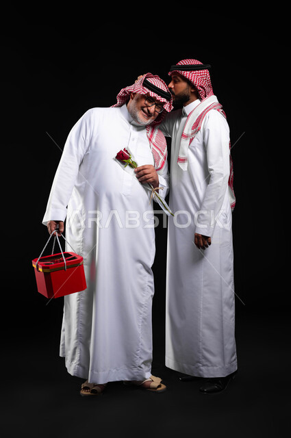 احترام وتقدير الآباء والإحسان لهم، إهداء الهدايا والزهور في الأعياد والمناسبات، بورتريه لشاب عربي خليجي سعودي يرتدي الثوب التقليدي والشماغ يقوم بتقبيل رأس والده، مفهوم المحبة والترابط الأسري، التربية الصالحة للأبناء، الطول الكامل للجسم، خلفية سوداء