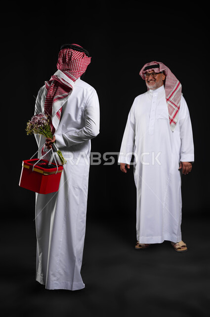 Gestures of joy and happiness in the presence of the son, surprising the father with his birthday gift, portrait of a Saudi Gulf Arab young man wearing a traditional dress and shemagh, hiding a red box and a bouquet of roses behind him, happy family celebrations and holidays, understanding of family intimacy and love, full length of the body, black background