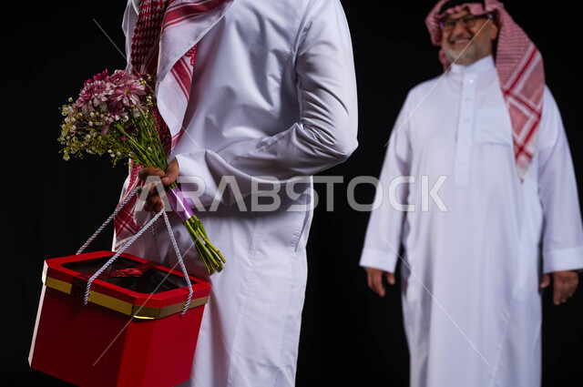 احتفالات وأعياد عائلية سعيدة، ايماءات البهجة والسرور بحضور الابن، مفاجأة الوالد بهدية عيد ميلاده، بورتريه مقرب من الخلف لشاب عربي خليجي سعودي يرتدي الزي التقليدي والشماغ يخبئ خلفه صندوق أحمر وباقة ورد، مفهوم الألفة والمحبة الأسرية، خلفية سوداء