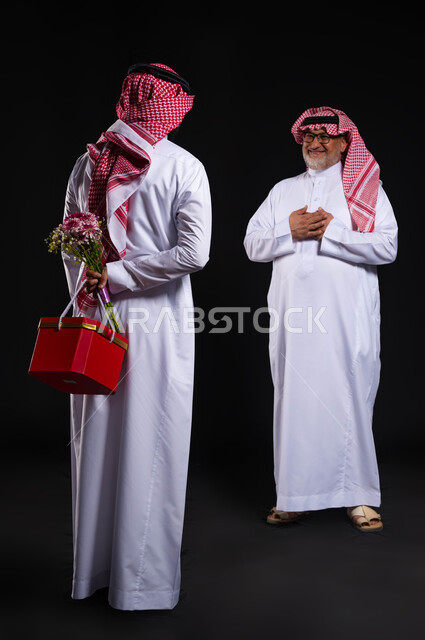 ايماءات تدل على الرضى والإمتنان، إدخال الفرحة والسرور الى قلب الأ ومفاجأته، بورتريه من الخلف لشاب عربي خليجي سعودي يرتدي الزي التقليدي والشماغ يحمل خلف ظهره باقة ورد وهدية، تقديم الهدايا في الأعياد والمناسبات السعيدة، الطول الكامل للجسم، خلفية سوداء