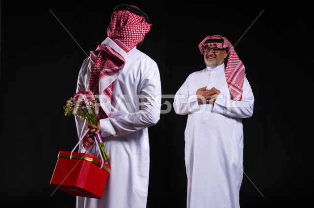 تقديم الهدايا في الأعياد والمناسبات السعيدة، إدخال الفرحة والسرور الى قلب الأب ومفاجأته، بورتريه من الخلف لشاب عربي خليجي سعودي يرتدي الزي التقليدي والشماغ يحمل خلف ظهره باقة ورد وهدية, ايماءات تدل على الرضى والإمتنان، خلفية سوداء