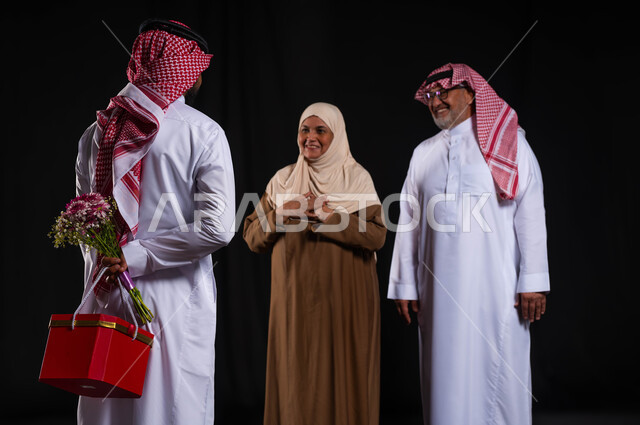 ايماءات السعادة والسرور، مفهوم رضى الوالدين والإحسان اليهم، أجواء عائلية سعيدة مليئة بالحب، بورتريه من الخلف لشاب عربي خليجي سعودي يرتدي الزي التقليدي والشماغ يحمل خلفه باقة ورد وصندوق هدية أحمر، الأعياد والمناسبات الأسرية السعيدة، خلفية سوداء