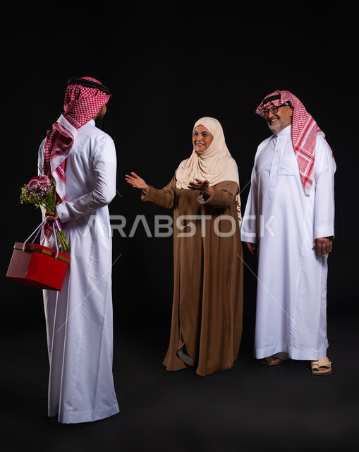 رضى الوالدين والإحسان اليهم، أجواء عائلية سعيدة مليئة بالحب، بورتريه من الخلف لشاب عربي خليجي سعودي يرتدي الزي التقليدي والشماغ يحمل خلفه باقة ورد وصندوق هدية أحمر، الأعياد والمناسبات الأسرية السعيدة، ايماءات السعادة والسرور، الطول الكامل للجسم، خلفية سوداء
