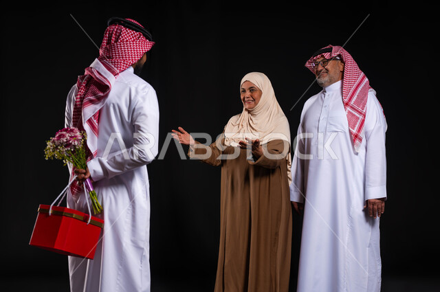 مفهوم رضى الوالدين والإحسان اليهم، أجواء عائلية سعيدة مليئة بالحب، بورتريه من الخلف لشاب عربي خليجي سعودي يرتدي الزي التقليدي والشماغ يحمل خلفه باقة ورد وصندوق هدية أحمر، الأعياد والمناسبات الأسرية السعيدة، ايماءات السعادة والسرور، خلفية سوداء
