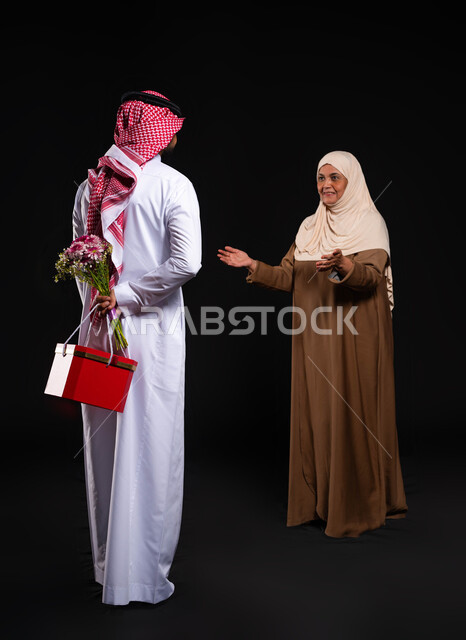 ايماءات تدل على البهجة والسرور في يوم الأم، مفهوم الأمومة والعطف والحنان، بورتريه من الخلف لشاب عربي خليجي سعودي يرتدي الثوب التقليدي يخبئ خلفه هدية وباقة ورد، إحياء ذكرى عيد ميلاد الوالدة بالهدايا والمفاجآت، الطول الكامل للجسم، خلفية سوداء