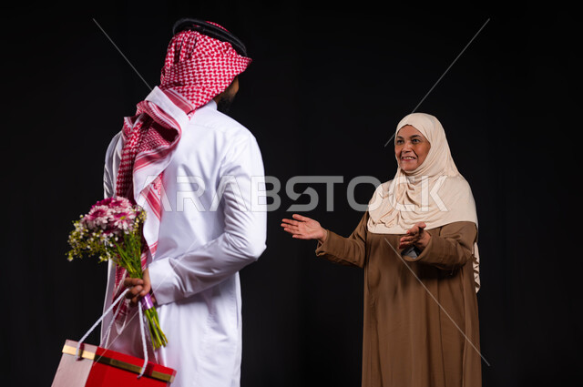 إحياء ذكرى عيد ميلاد الوالدة بالهدايا والمفاجآت، مفهوم الأمومة والعطف والحنان، بورتريه من الخلف لشاب عربي خليجي سعودي يرتدي الثوب التقليدي يخبئ خلفه هدية وباقة ورد، ايماءات تدل على البهجة والسرور في يوم الأم، خلفية سوداء