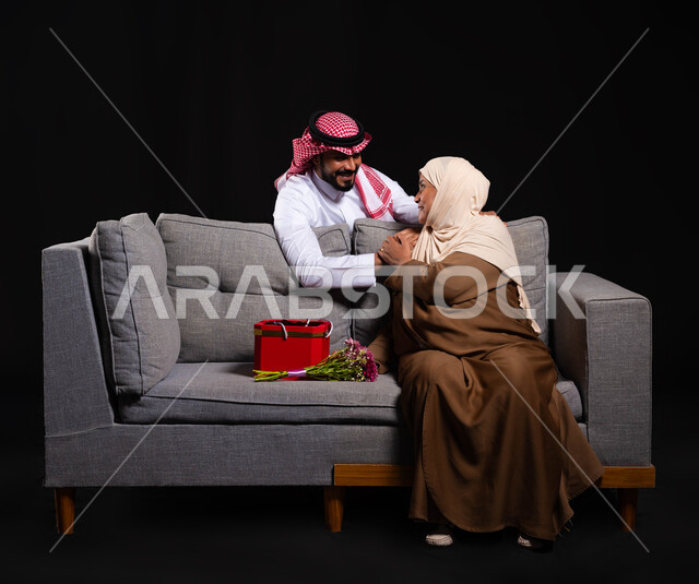 تقدير وحب الأم والإحسان إليها، ايماءات الرضى والمودة، بورتريه لشاب عربي خليجي سعودي يرتدي الثوب التقليدي والشماغ يقف خلف الأريكة التي تجلس عليها والدته، تقديم الهدايا والأزهار للأمهات بذكرى عيد الأم، الاحتفال بالأعياد والمناسبات، الطول الكامل للجسم، خلفية سوداء