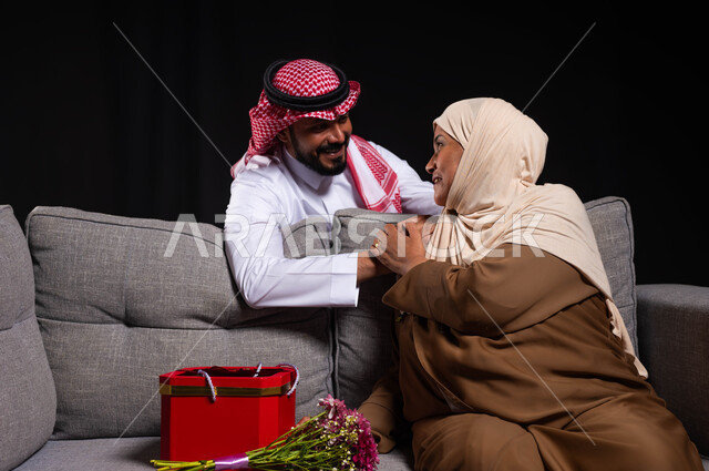 حب وتقدير الأم والإحسان إليها، ايماءات الرضى والمودة، بورتريه لشاب عربي خليجي سعودي يرتدي الثوب التقليدي والشماغ يقف خلف الأريكة التي تجلس عليها والدته، تقديم الهدايا والأزهار للأمهات بذكرى عيد الأم، الاحتفال بالأعياد والمناسبات العائلية، خلفية سوداء