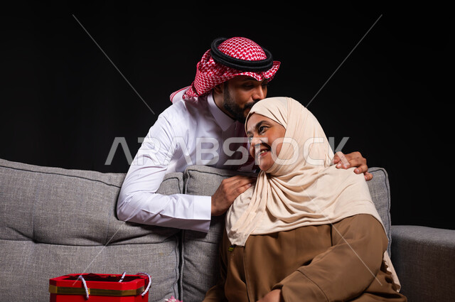 ايماءات الرضى والسرور، مفهوم حب الأم والإحسان إليها، بورتريه مقرب لشاب عربي خليجي سعودي يرتدي الثوب التقليدي والشماغ يقوم بتقبيل رأس أمه واحضار هدية لها، تقديم الهدايا والأزهار للأمهات بذكرى عيد الأم، الاحتفال بالأعياد والمناسبات العائلية، خلفية سوداء