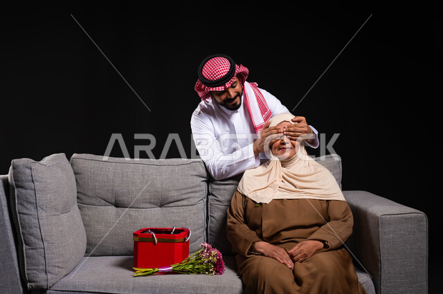 الاحتفال بالأعياد والمناسبات السعيدة، بورتريه لشاب عربي خليجي سعودي يرتدي الثوب التقليدي والشماغ يغمضعيني والدته بيده، مفاجأة الأم وإدخال السرور عليها، مفهوم بر الوالدين ورضا الأمهات، التربية الصالحة للأبناء، أجواء عائلية مليئة بالحب، خلفية سوداء