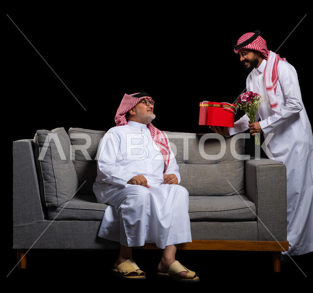 احتفالات وأعياد وأجواء سعيدة، التعبير عن المحبة والألفة في المناسبات، تقديم الهدايا التذكارية للآباء، بورتريه لشاب عربي خليجي سعودي يرتدي الزي التقليدي والشماغ يقوم بتقديم باقة ورد وهدية لوالده، ايماءات الفرح والسعادة والمحبة، الطول الكامل للجسم، خلفية سوداء