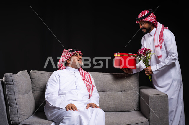 التعبير عن المحبة والألفة في المناسبات، تقديم الهدايا التذكارية للآباء، بورتريه لشاب عربي خليجي سعودي يرتدي الزي التقليدي والشماغ يقوم بتقديم باقة ورد وهدية لوالده، ايماءات الفرح والسعادة والمحبة، أعياد واحتفالات وأجواء سعيدة، خلفية سوداء