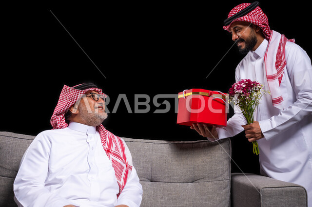 تقديم الهدايا التذكارية للآباء، التعبير عن المحبة والألفة في المناسبات، بورتريه لشاب عربي خليجي سعودي يرتدي الزي التقليدي والشماغ يقوم بتقديم باقة ورد وهدية لوالده، ايماءات الفرح والسعادة والمحبة، أعياد واحتفالات وأجواء سعيدة، خلفية سوداء